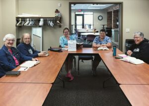 An image of 5 women seated in a conference room. They are smiling and laughing, and have note taking materials and bibles in front of them.